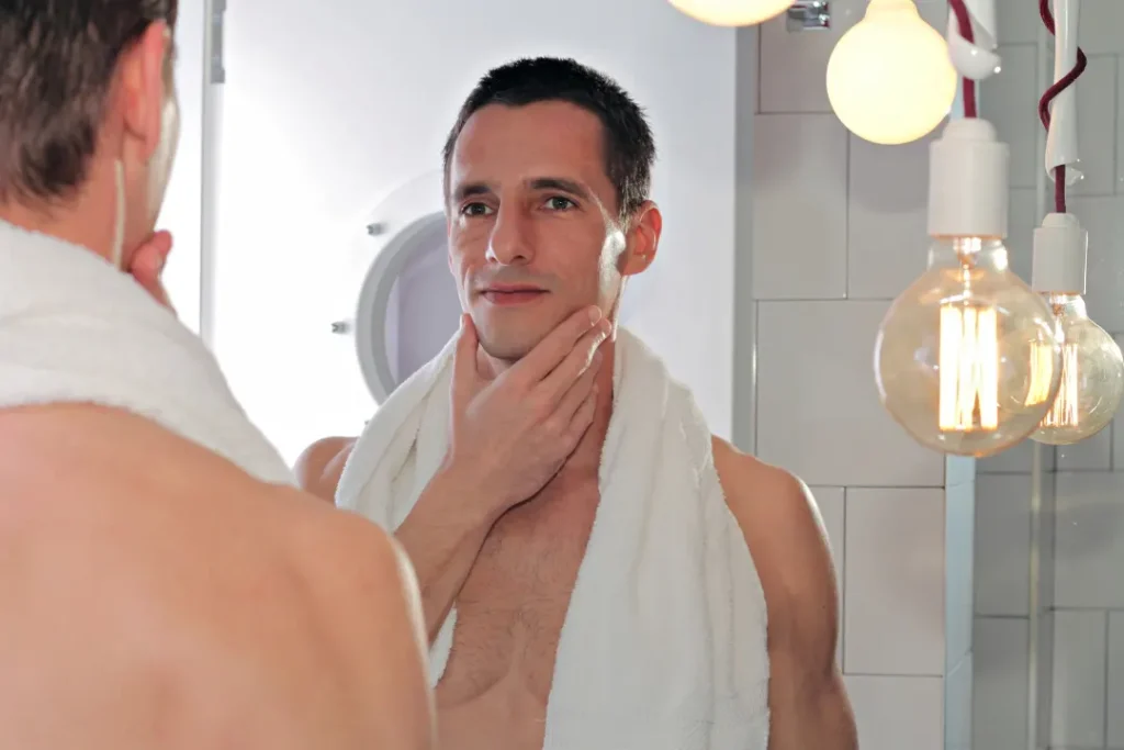 A young attractive man touches his face skin while standing in front of the mirror. 