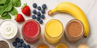 Four glass jars of colorful healthy smoothies surrounded by fresh ingredients like berries, spinach, chia seeds, bananas, nut butters, yogurt, and protein powder on a marble surface.